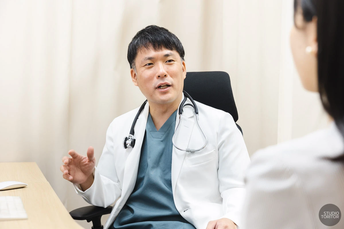 内科クリニックの診察室で医師が患者に丁寧に説明を行う診療風景写真。ホームページ用撮影事例。
