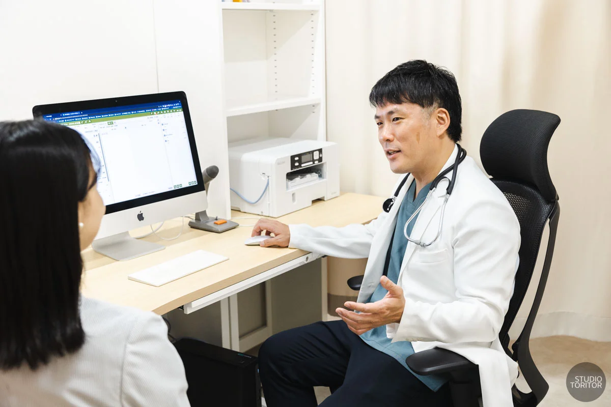 電子カルテを見ながら患者にわかりやすく説明する医師の診察風景。クリニック出張撮影事例。