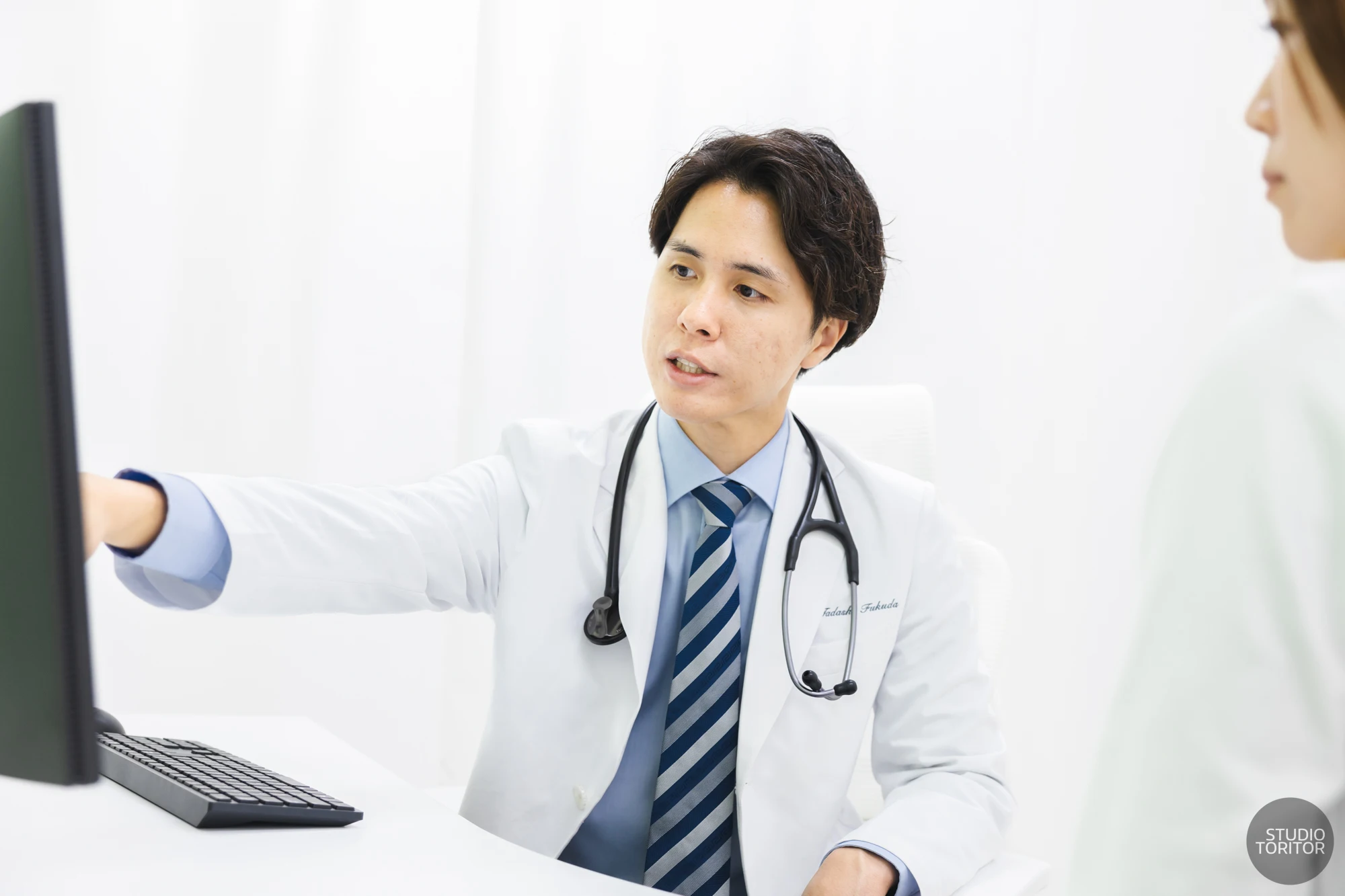 内科・循環器内科クリニックの医師が診察室でPC画面を指さし患者に説明する様子。ホームページ用写真撮影事例。