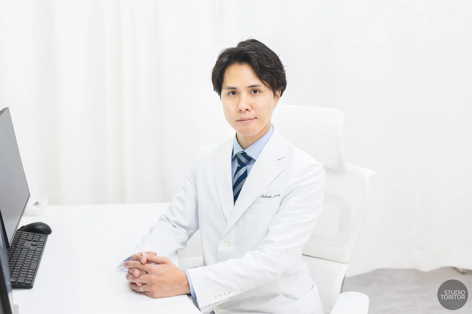 内科クリニックの医師(院長)が診察室デスクで真剣な表情で座るポートレート写真。採用サイト・スタッフ紹介向け。