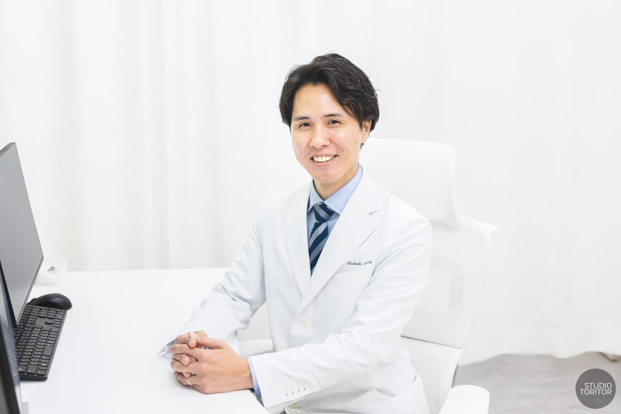 内科・循環器内科診療所の医師(院長)が診察室デスクで笑顔で座る写真。スタッフ紹介用。