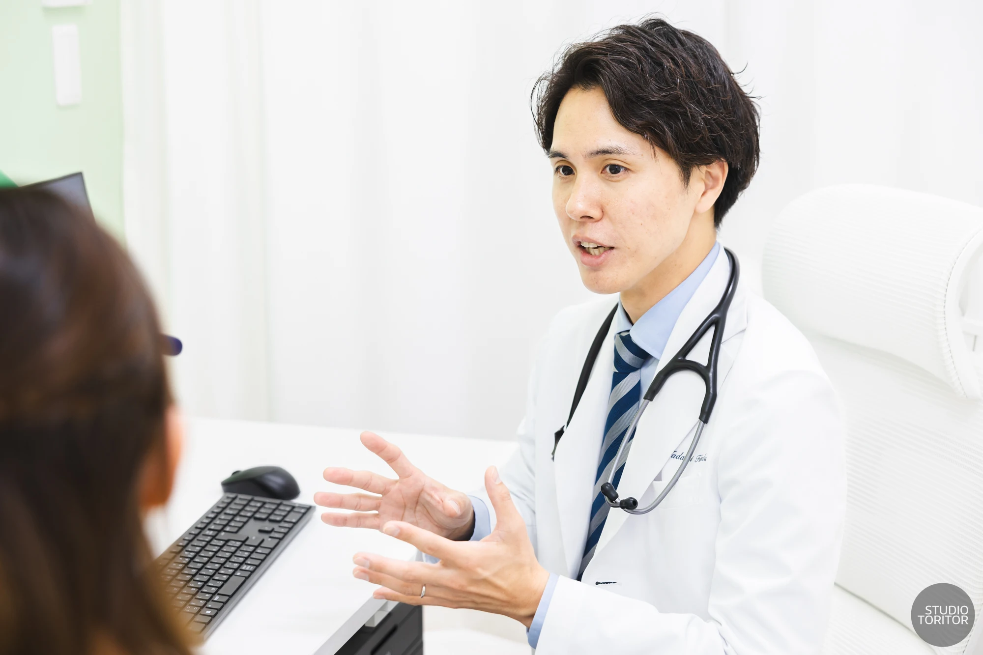 内科クリニックの医師(院長)が診察室で患者にジェスチャーを交えて真剣に説明する様子。業務風景写真。