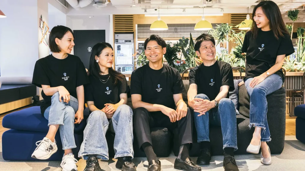 ロゴ入りTシャツを着たスタッフ5人がソファーで談笑する社内風景（企業のスタッフ採用・ブランディング写真撮影実績）