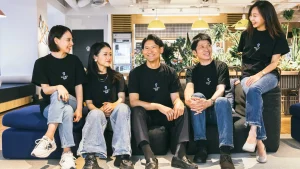 ロゴ入りTシャツを着たスタッフ5人がソファーで談笑する社内風景（企業のスタッフ採用・ブランディング写真撮影実績）