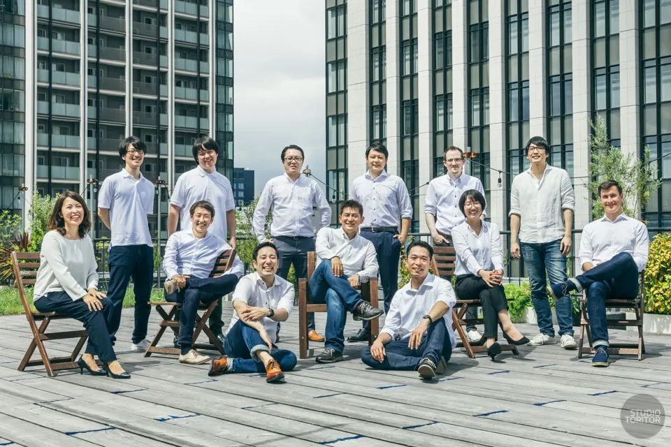 開放的な屋外ウッドデッキで撮影したチームの一体感と爽やかさを伝える企業集合写真